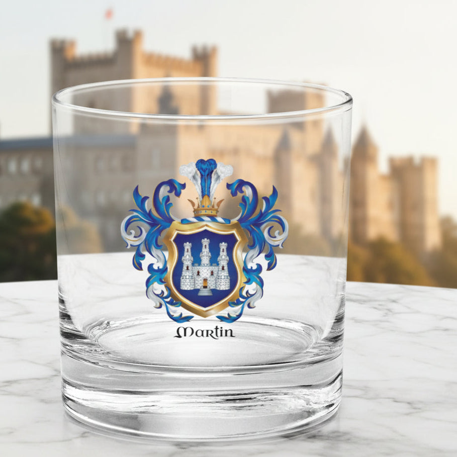 A clear glass rocks tumbler featuring a printed Martin family crest with blue shield and white castle, set against a background with a view of a castle