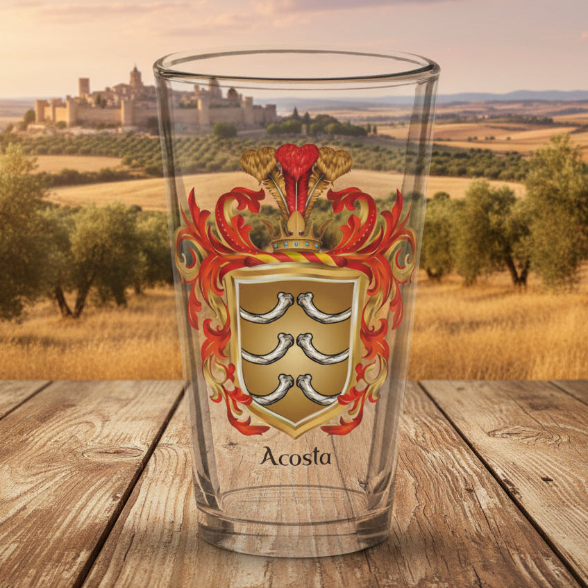A clear pint glass featuring a printed Acosta family crest with red, gold, and silver details, and the name 'Acosta' printed below in black, shown on an old wooden tabletop with a rural Spanish landscape with a walled Spanish city in the distance.
