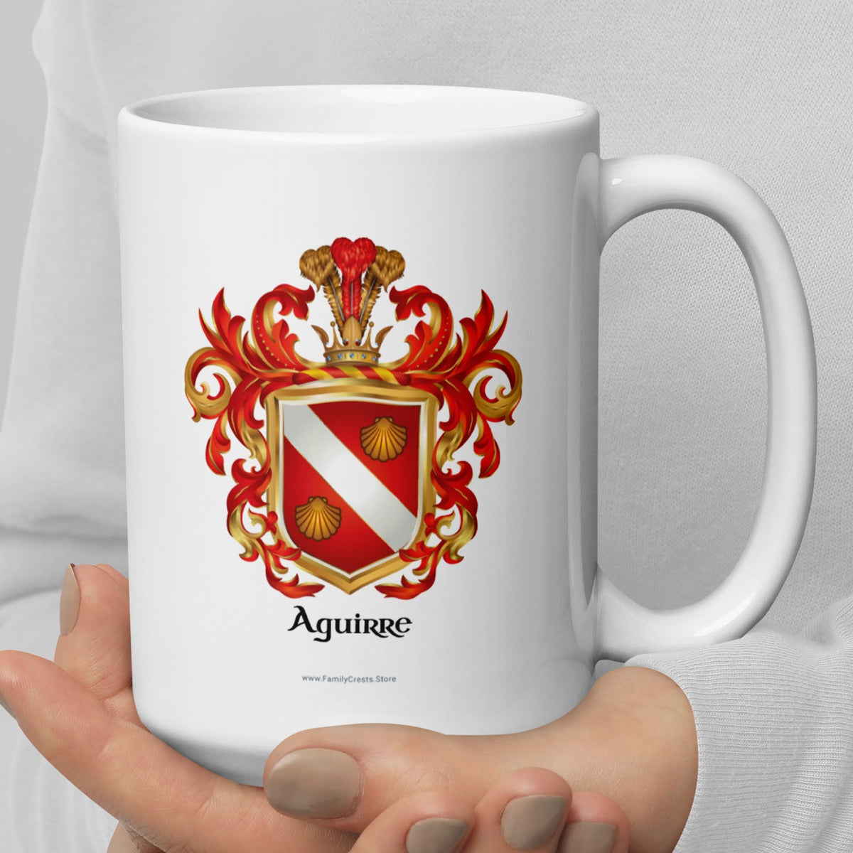 A person in a white long-sleeve top holds a white ceramic mug with a red, gold, and white Aguirre coat of arms printed on the front, set against a neutral light gray background.