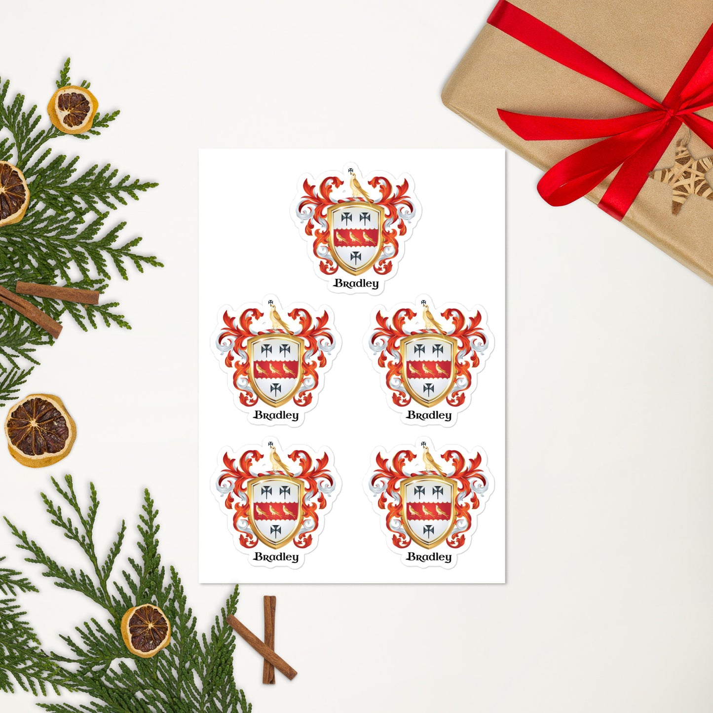 A printed sheet showing five identical coat of arms-style crest designs labeled 'Bespley,' arranged on a white background with holiday greenery, dried orange slices, cinnamon sticks, and a wrapped gift box with a red ribbon nearby.