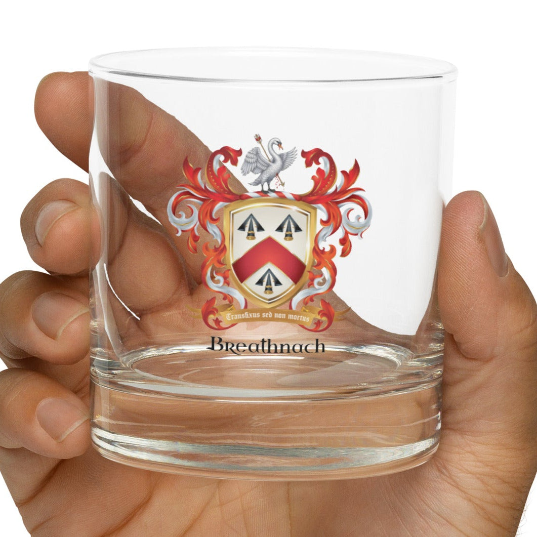 A hand holding a clear glass rocks tumbler featuring a printed family crest with red, white, and black details, and the name 'Breathnach' below the shield.