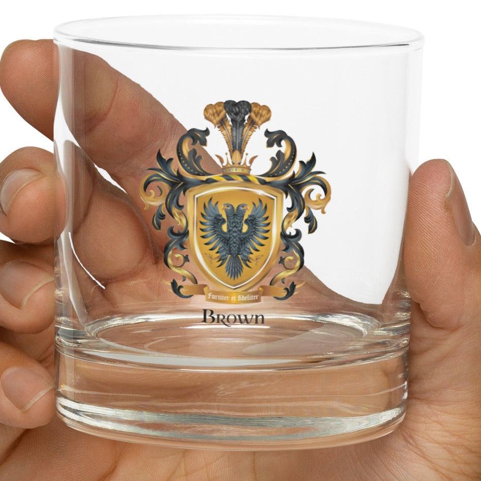 Hand holding a clear rocks glass with an illustration of the Brown Family Crest with 2 headed eagle on a white background