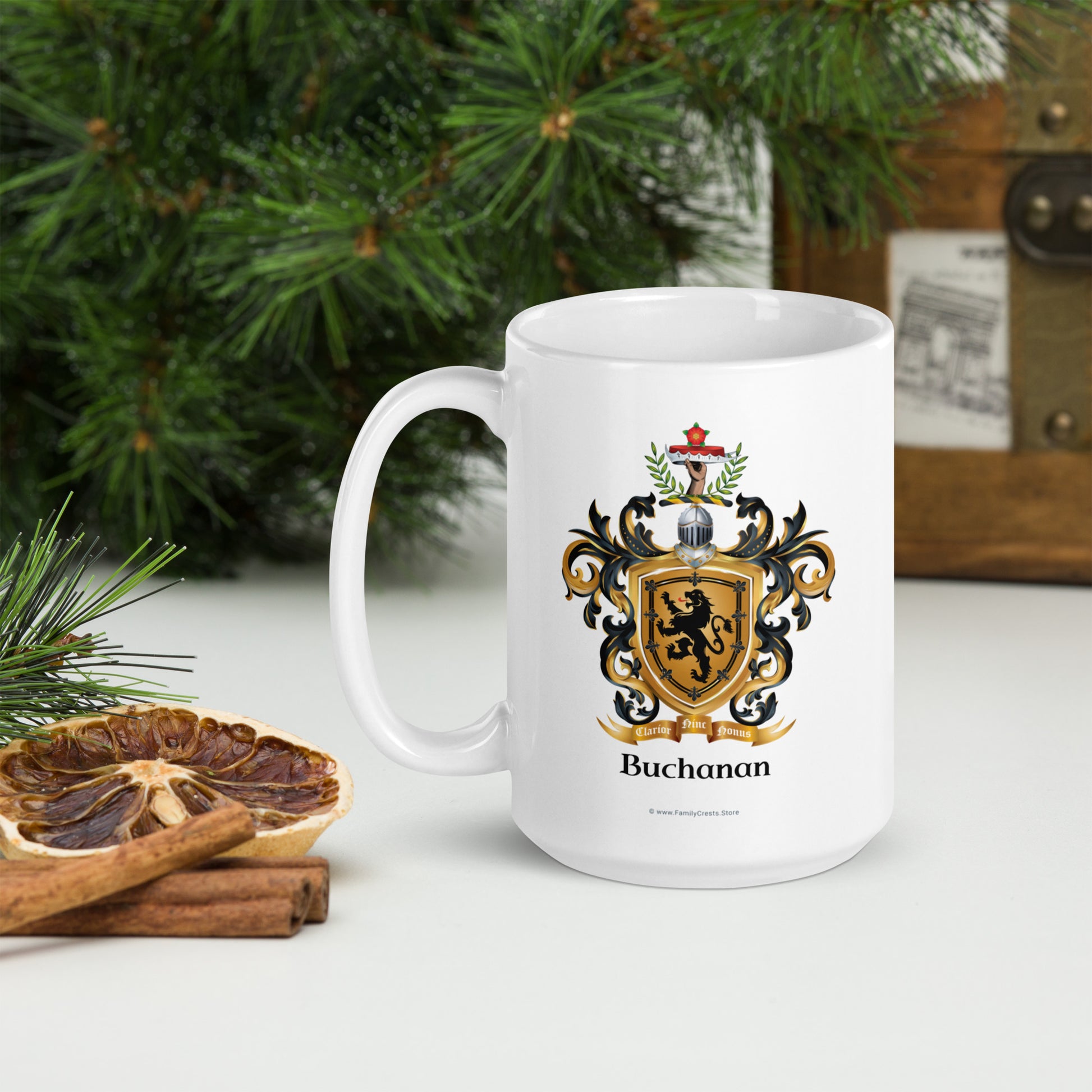 A white ceramic mug with a printed Buchanan family crest and the name 'Buchanan' below it, styled on a tabletop with pine branches, a dried orange slice, cinnamon sticks, and a wooden chest in the background.