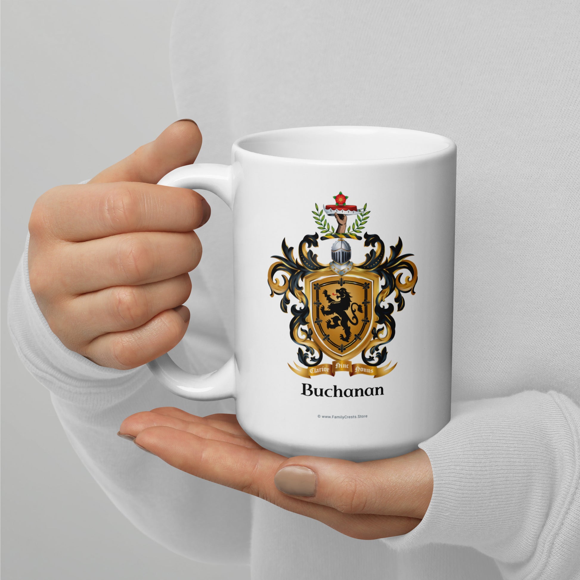 A white ceramic mug with a family crest and the name 'Buchanan' printed on it, held by a person's hands against a light gray background.
