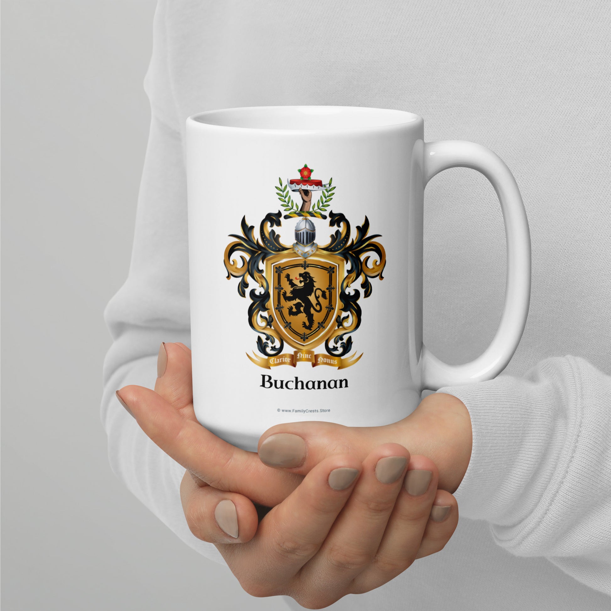 A person in a white long-sleeve top holds a white ceramic mug with a detailed Buchanan family crest printed on the front. The crest features a gold lion on a black and gold shield.