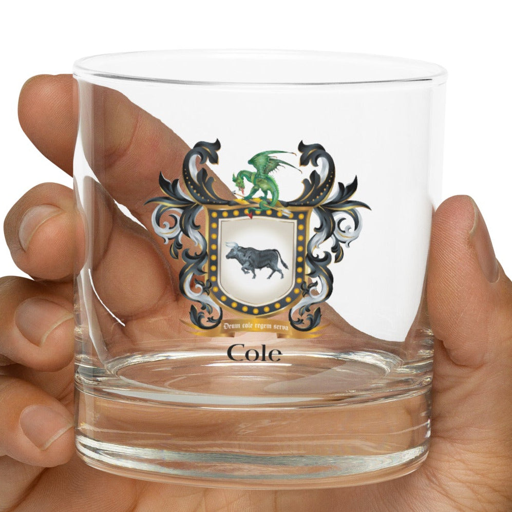Hand holding a clear glass with a Cole family crest coat of arms design on a white background 