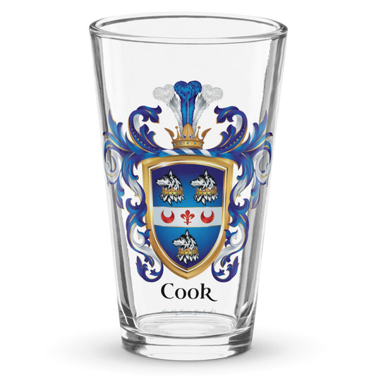 A clear classic pint beer glass on a white background, printed with the Cook Family Crest featuring a blue shield with 3 wolf heads with crowns around their necks and a white band with 2 red crescents and a fleur de lis.