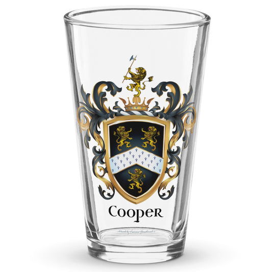 A clear classic pint beer glass on a white background, printed with the Cooper Family Crest featuring 3 lions and a patterned chevron on a black shield.