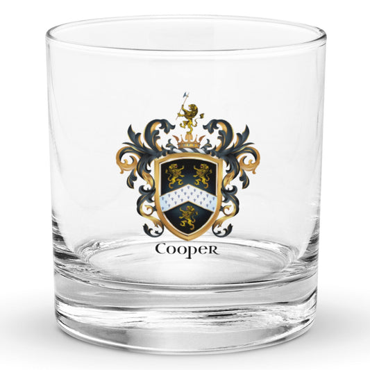 A clear classic heavy base rocks glass on a white background, printed with the Cooper Family Crest featuring 3 lions and a patterned chevron on a black shield.
