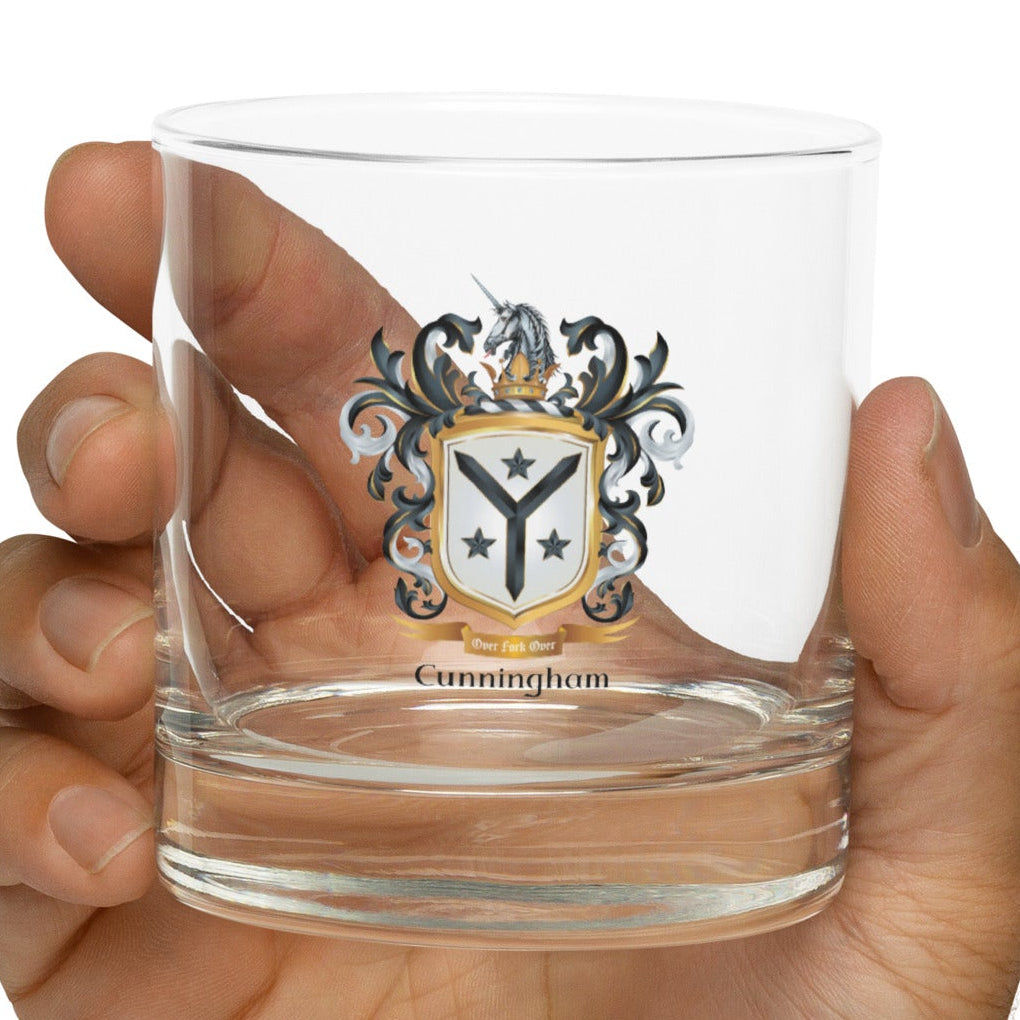 Hand holding a clear glass with a Cunningham family coat of arms design on a white background