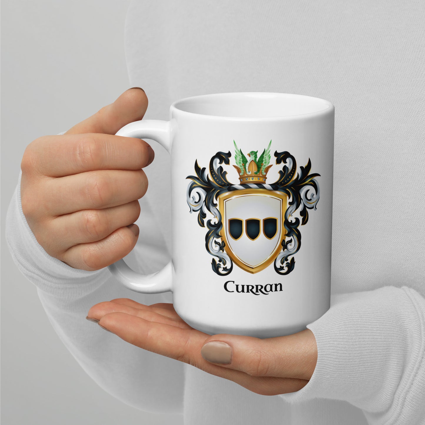A person in a white long-sleeve top holds a white ceramic mug with a printed family crest and the name 'Curran' below it. The mug is shown against a soft light gray background.
