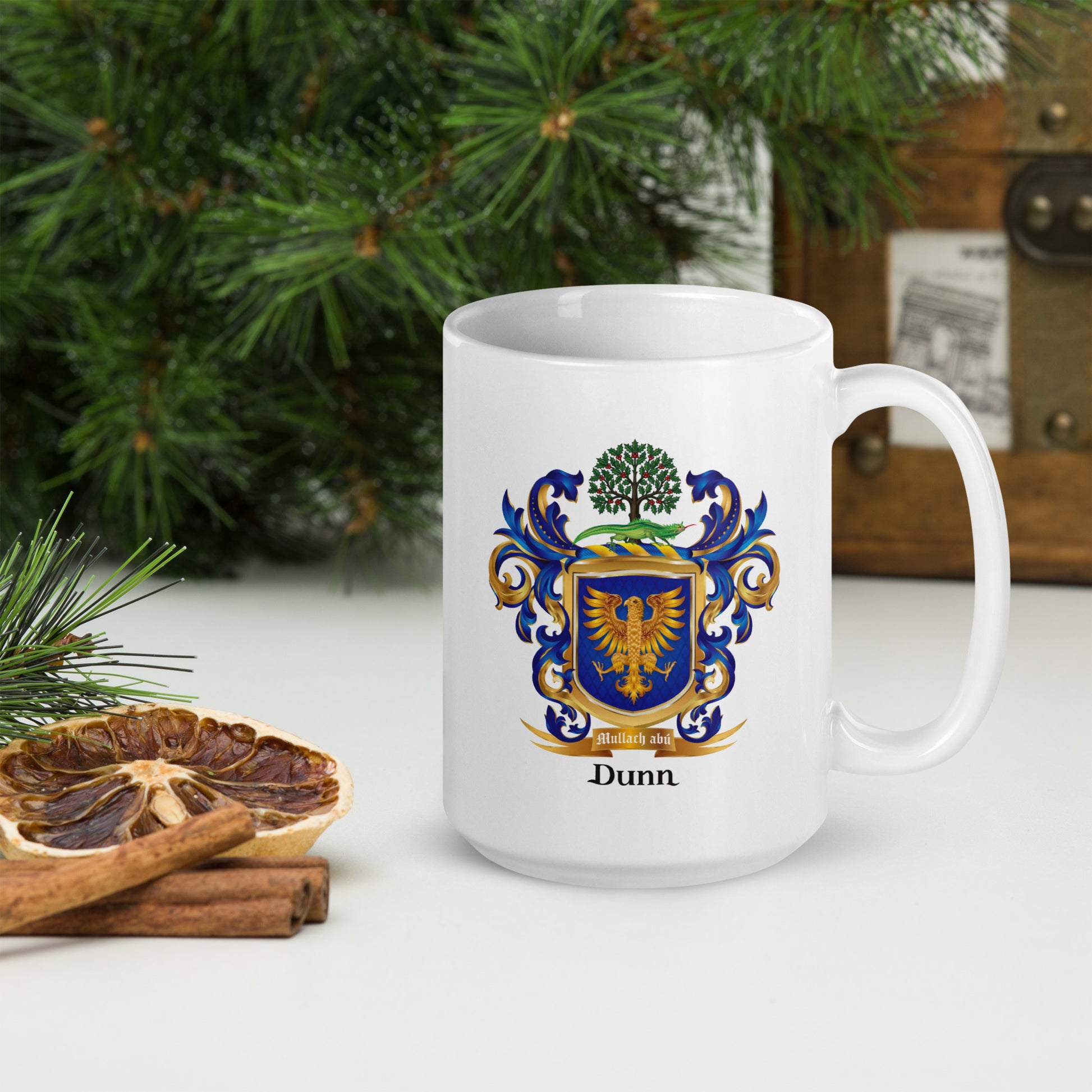 A white ceramic mug with a printed blue, gold, and green family crest and the name 'Dunn' below it, placed on a tabletop with pine branches, dried orange slices, and cinnamon sticks in a holiday setting.