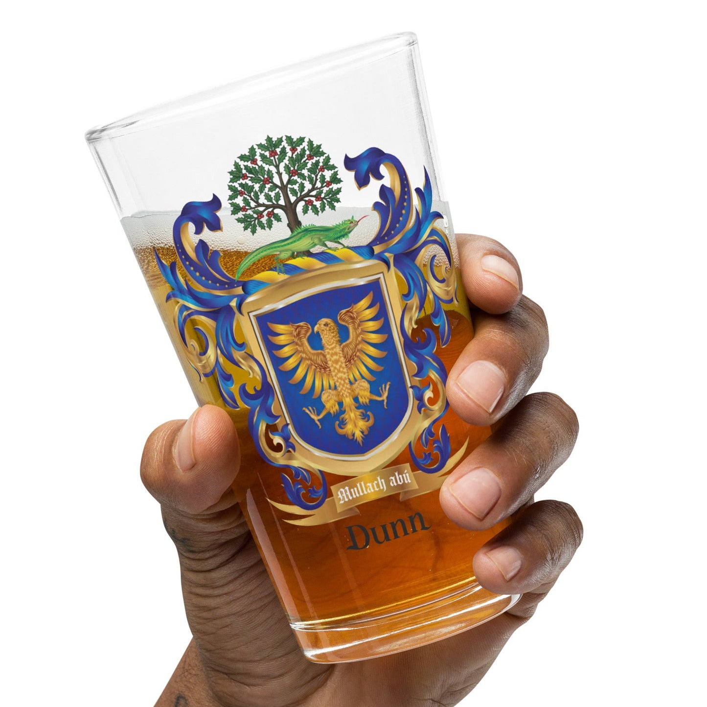 A hand holding a clear pint glass filled with amber beer, featuring a printed blue, gold, and green coat of arms crest with the name 'Dunn' near the bottom, against a plain white background.