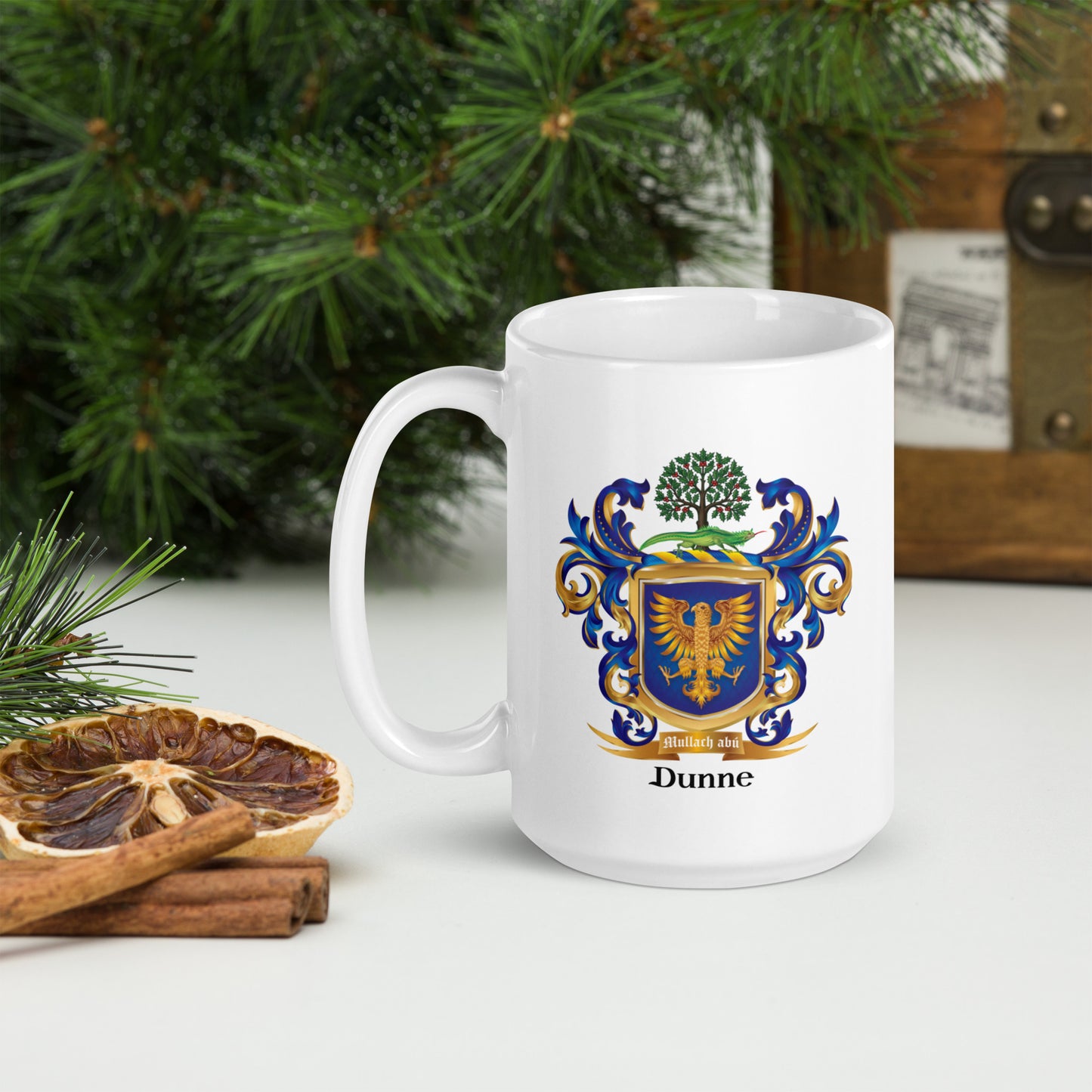 A white ceramic mug with a blue, gold, and green Dunne family crest printed on the front, set on a white table next to a dried orange slice, cinnamon sticks, and green pine branches.
