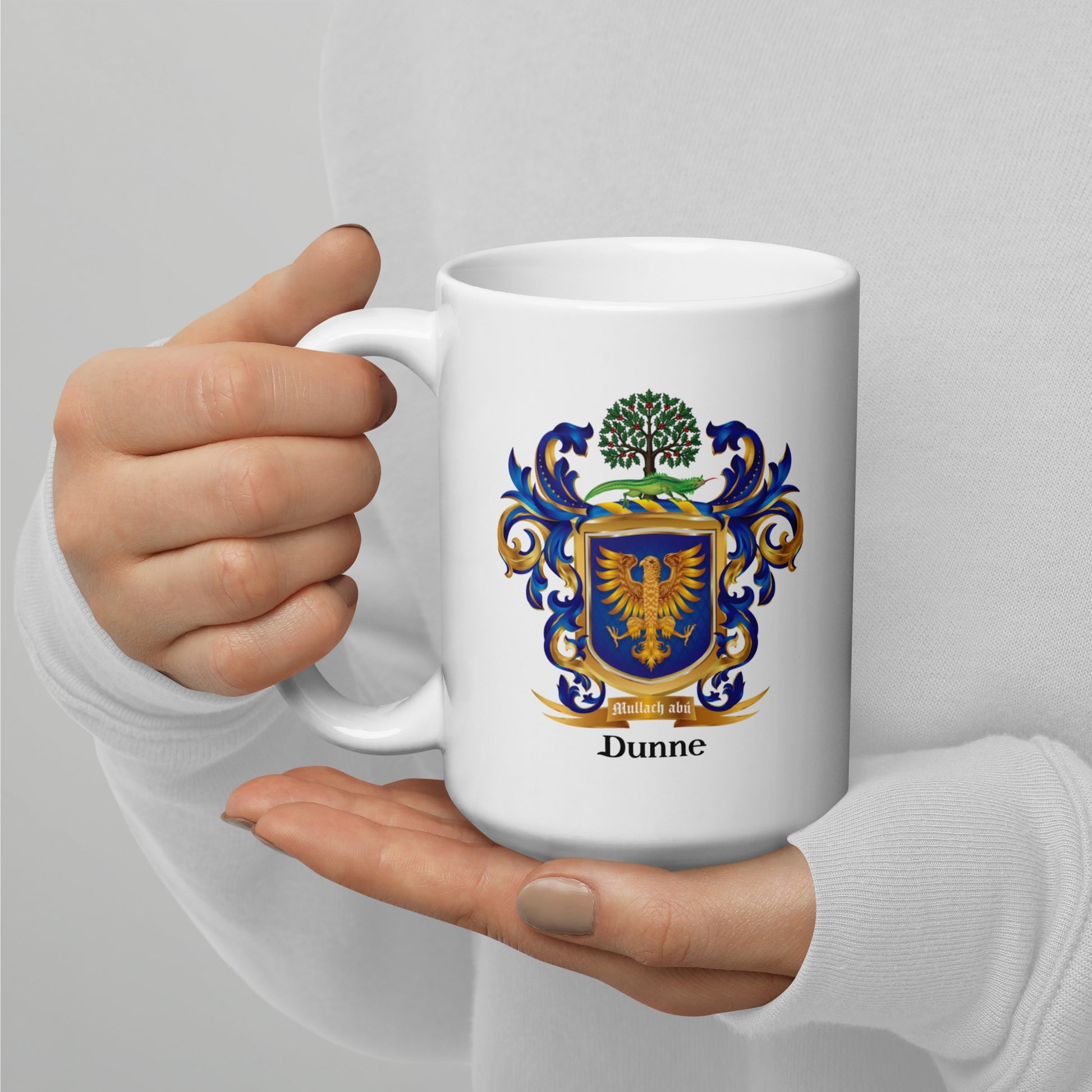 A person holding a white ceramic mug with a printed Dunne family crest in blue, gold, and green, against a light gray background.