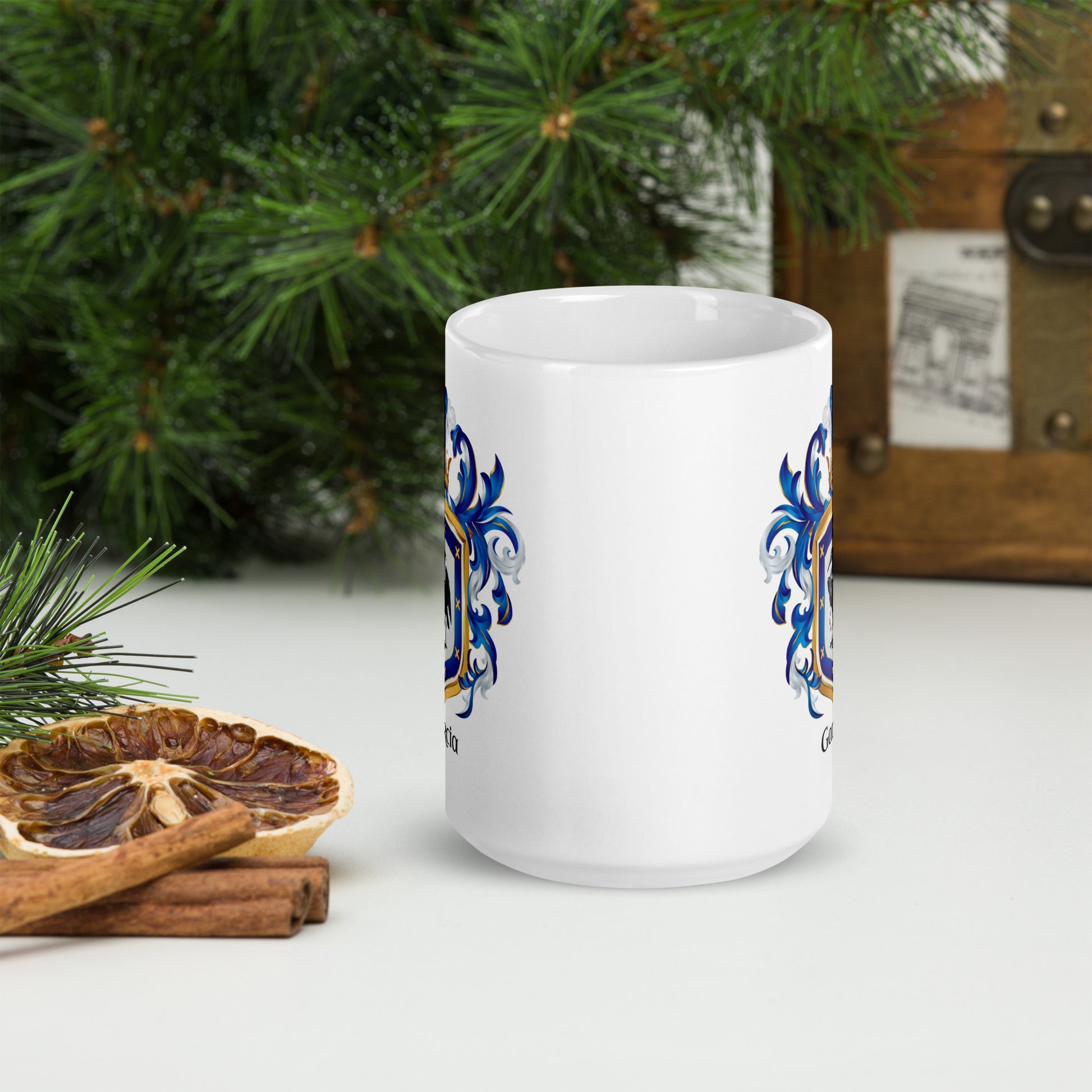 White mug with two Garcia blue and gold coat of arms on a white surface with Christmas decorations in the background
