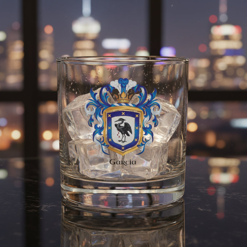 A clear rocks glass with ice cubes with a Garcia Family Crest on the front of the glass showing a heron on a white and blue shield with the name Garcia under the coat of armour in black text, on a marble table with city lights in the distant background visible through windows