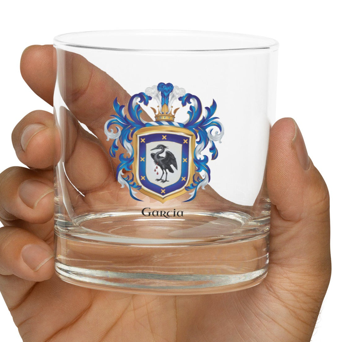 A clear rocks glass with a Garcia Family Crest showing a heron on a white and blue shield with the name Garcia under the coat of armour in black text, held in a hand against a white background
