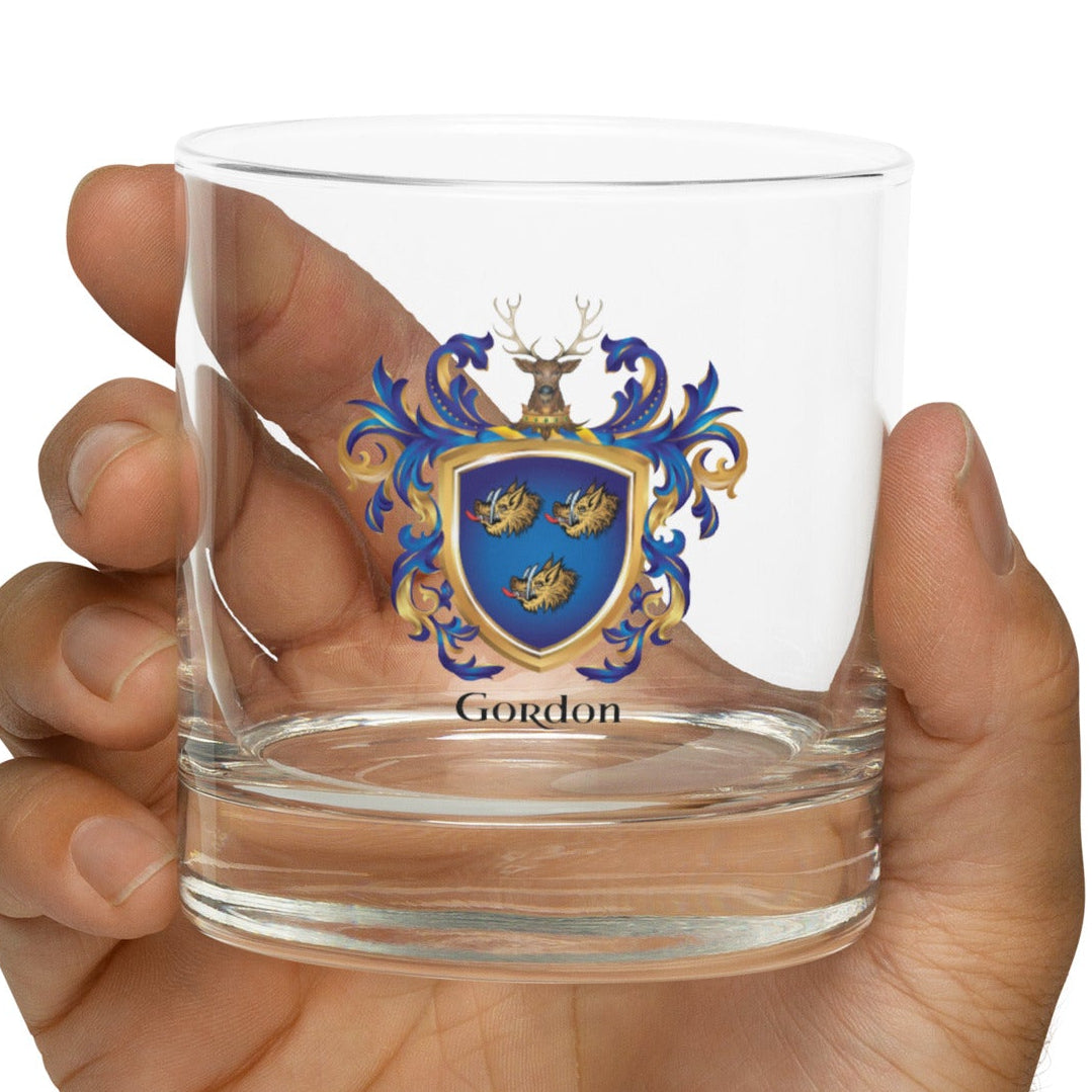 A hand holding a clear rocks glass with a Gordon family coat of arms crest and the name 'Gordon' printed below it, against a plain white background.