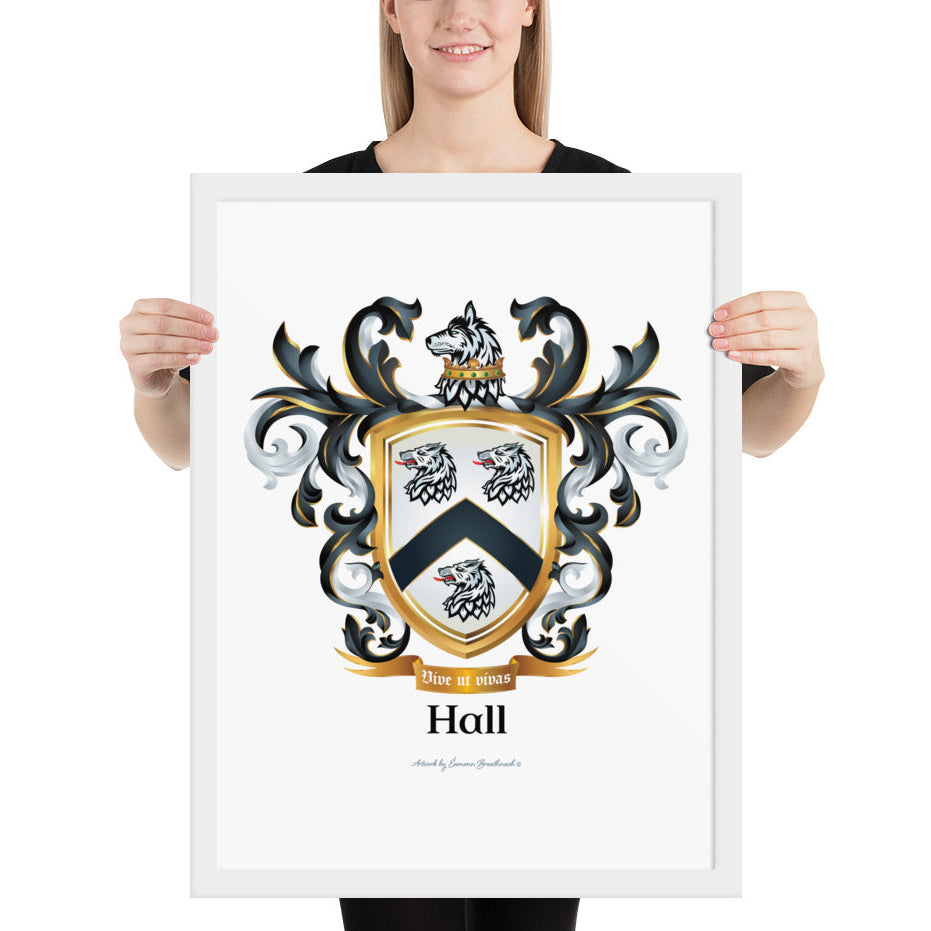 A woman standing in a white background, holding a white framed print of the Hall Family Crest. The crest features a white shield with a black chevron between three wolf heads (two above, one below), topped by wolf with a gold crown around it's neck and surrounded by detailed black and white mantling. The family name and motto appear beneath the shield.