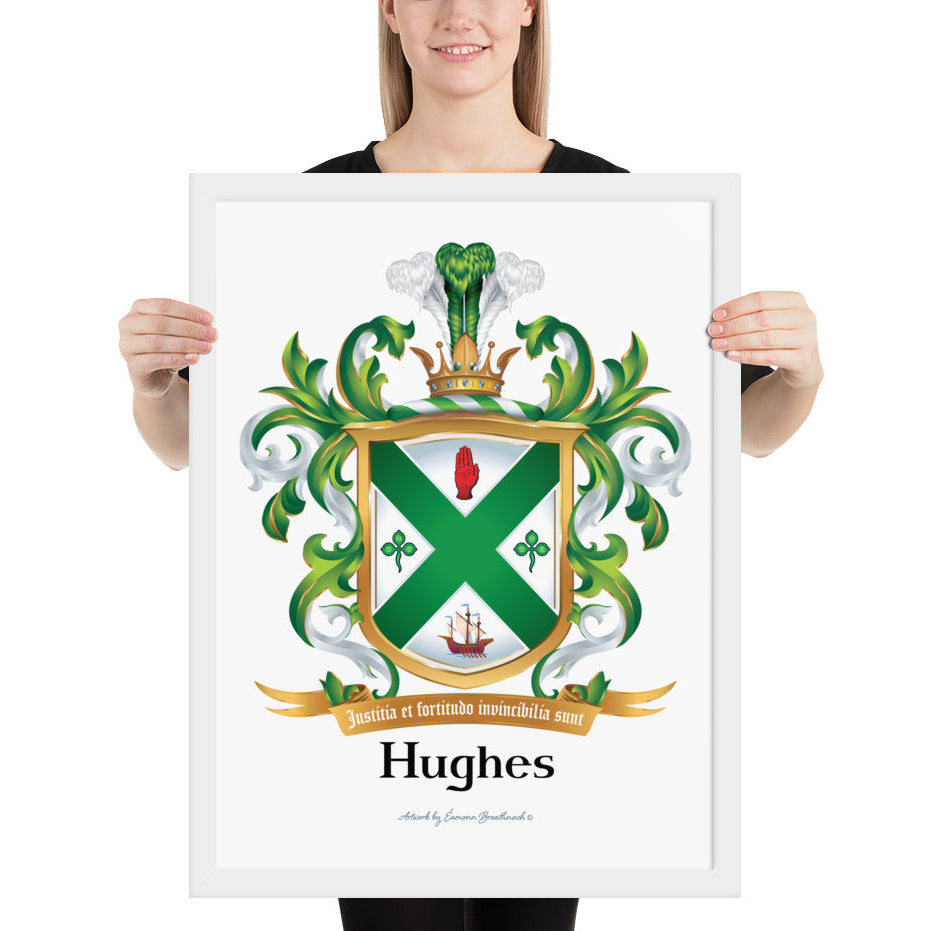 A woman standing in a white background, holding a white framed print of the Irish Hughes Family Coat of Arms featuring a green and white shield, a gold border, 2 green shamrocks, a s sailing ship, a gold motto scroll reading ‘Justitia et fortitudine invincibilia sunt’, and the name ‘Hughes’ below inheraldic style black text on a white background.