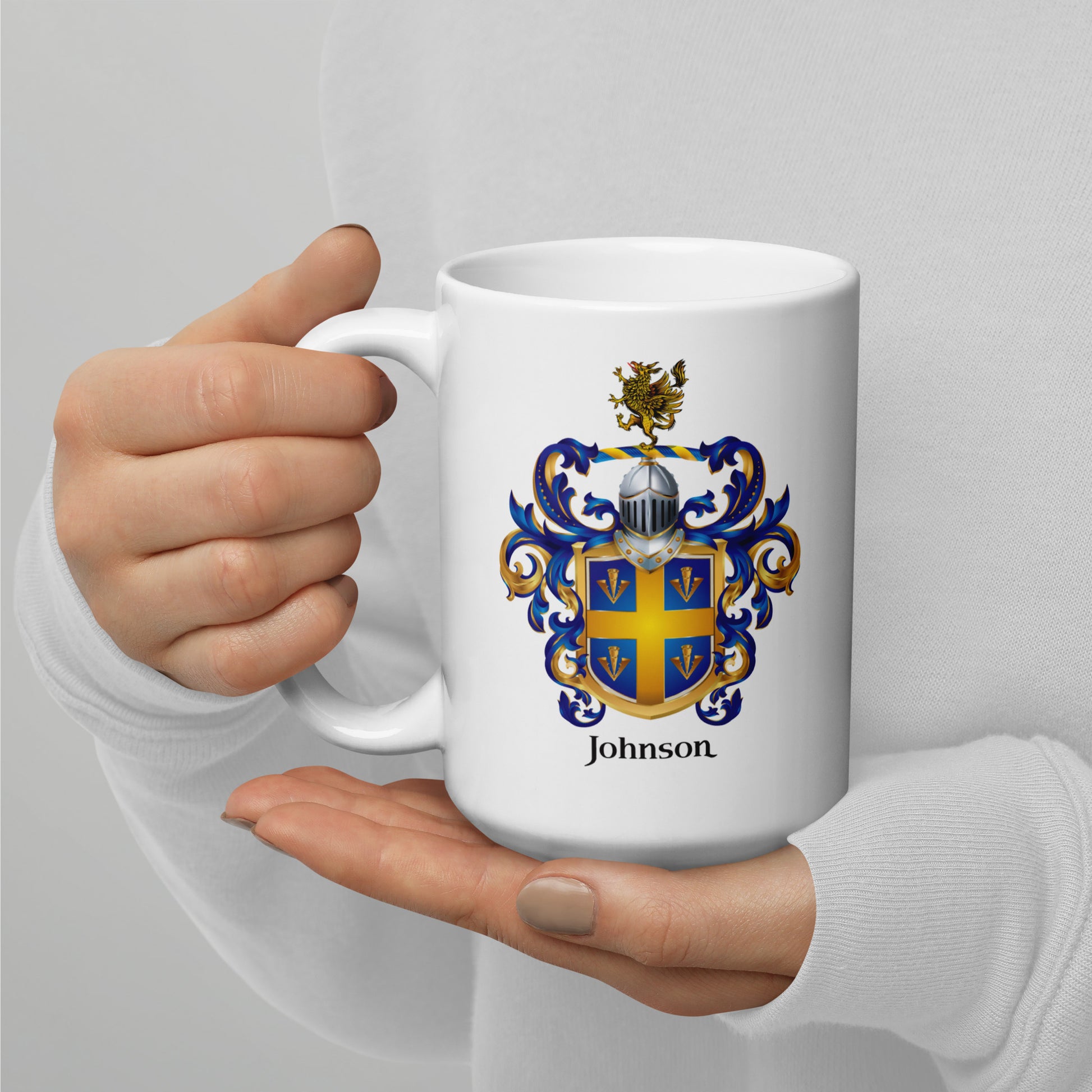 A person holding a white ceramic mug with a custom family crest and the name 'Johnson' printed on it. The mug features a blue and gold coat of arms with a silver helmet and crestal crest, and the name 'Johnson' is printed below in black.