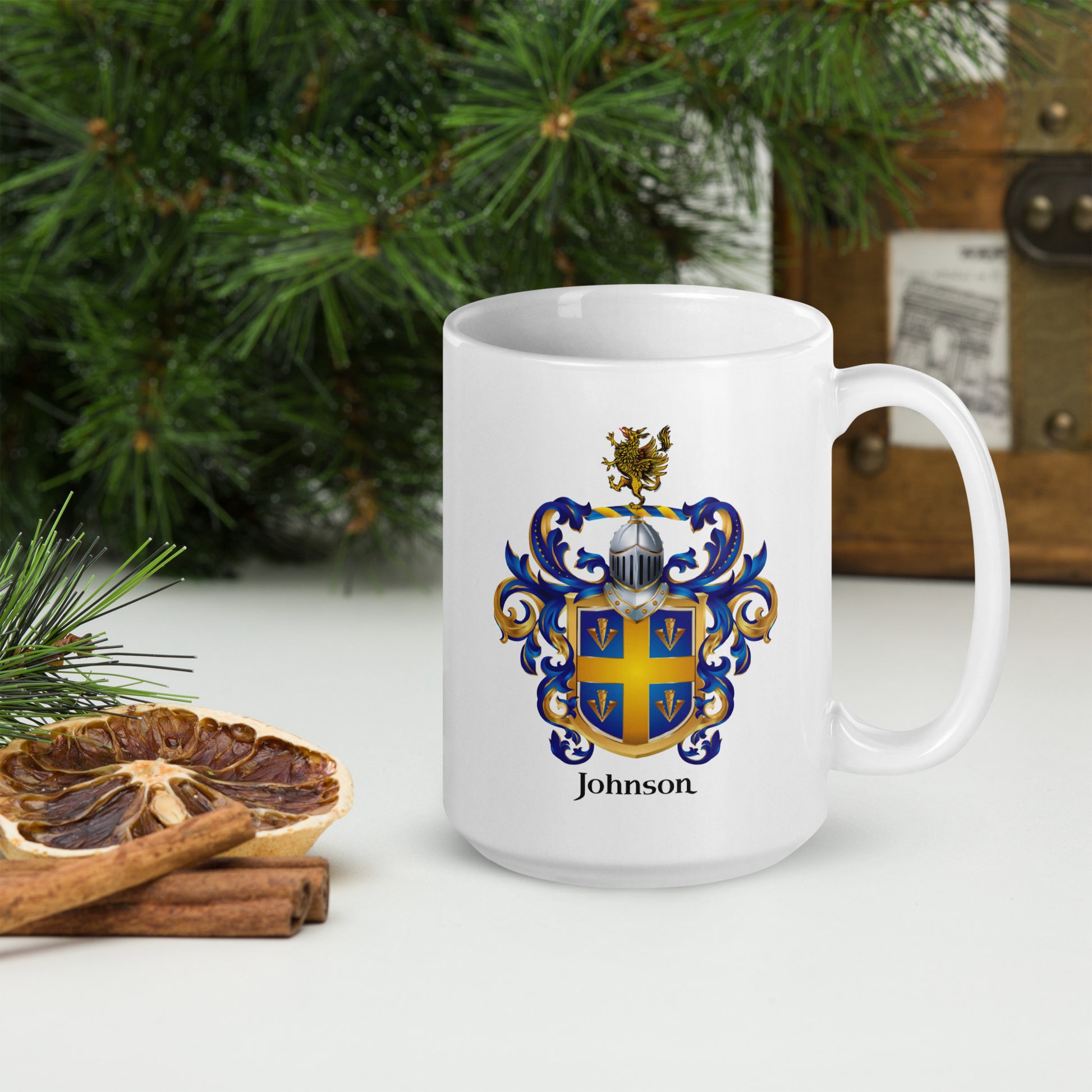 A white ceramic mug with a personalized family crest design featuring the name 'Johnson' and a blue, gold, and brown coat of arms. The mug is styled on a white surface with a dried orange slice, cinnamon sticks, and green pine branches in the background.
