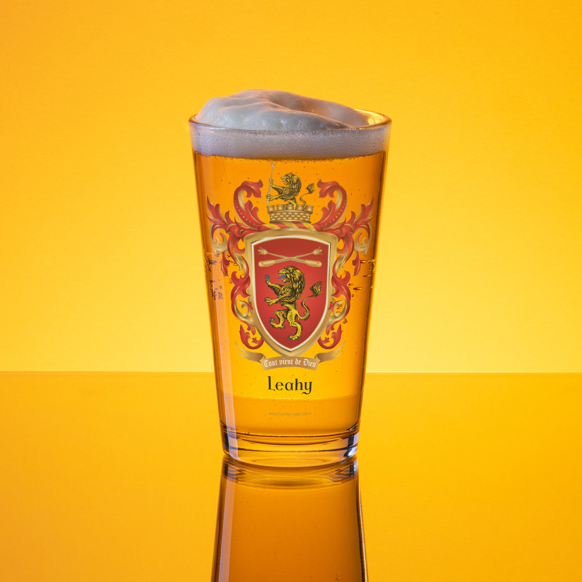 A clear pint glass filled with golden beer, topped with white foam, standing on a reflective yellow surface against a bright yellow background. The glass features a red and gold crest design with the name 'Leahy' printed near the bottom.