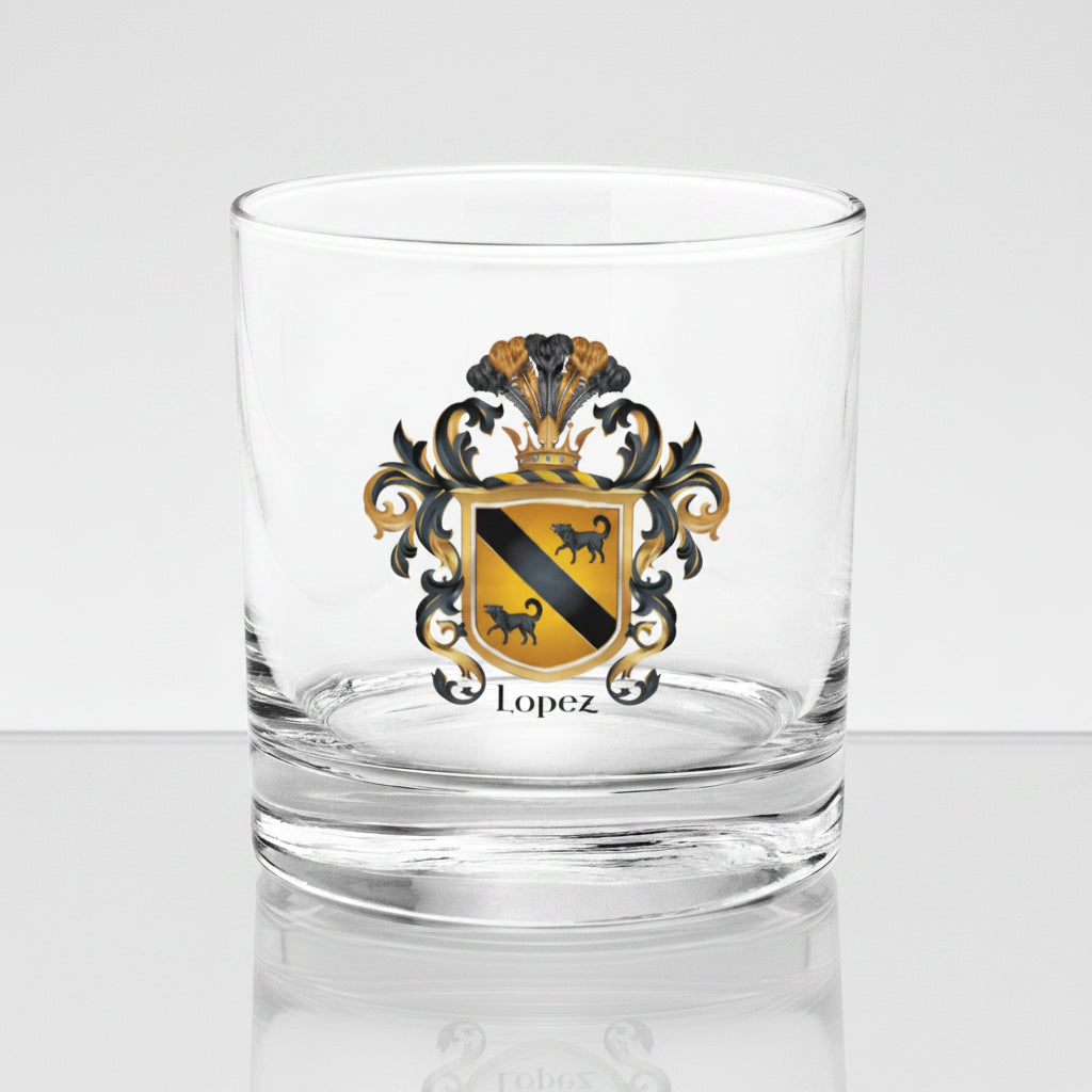 A rocks glass with the Lopez Family Crest printed on the front featuring a gold shield with a black diagonal band and two black wolves and the name Lopez in black text below the shield. The glass is sitting on a glass table.