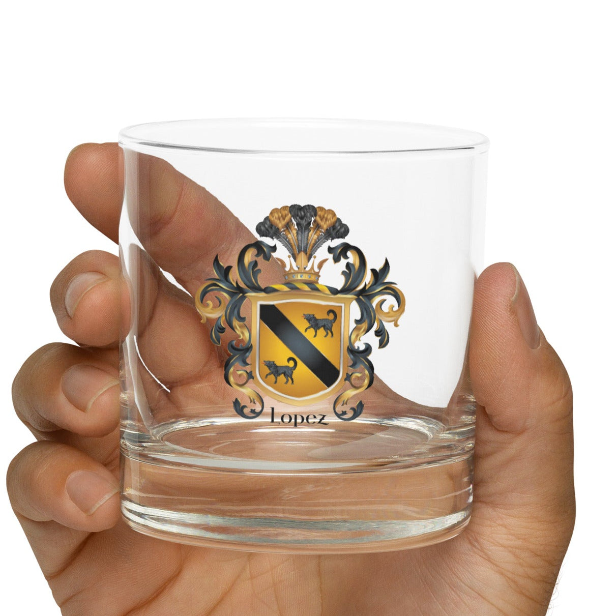 Hand holding arocks glass with the Lopez Family Crest printed on the front featuring a gold shield with a black diagonal band and two black wolves and the name Lopez in black text below the shield. 