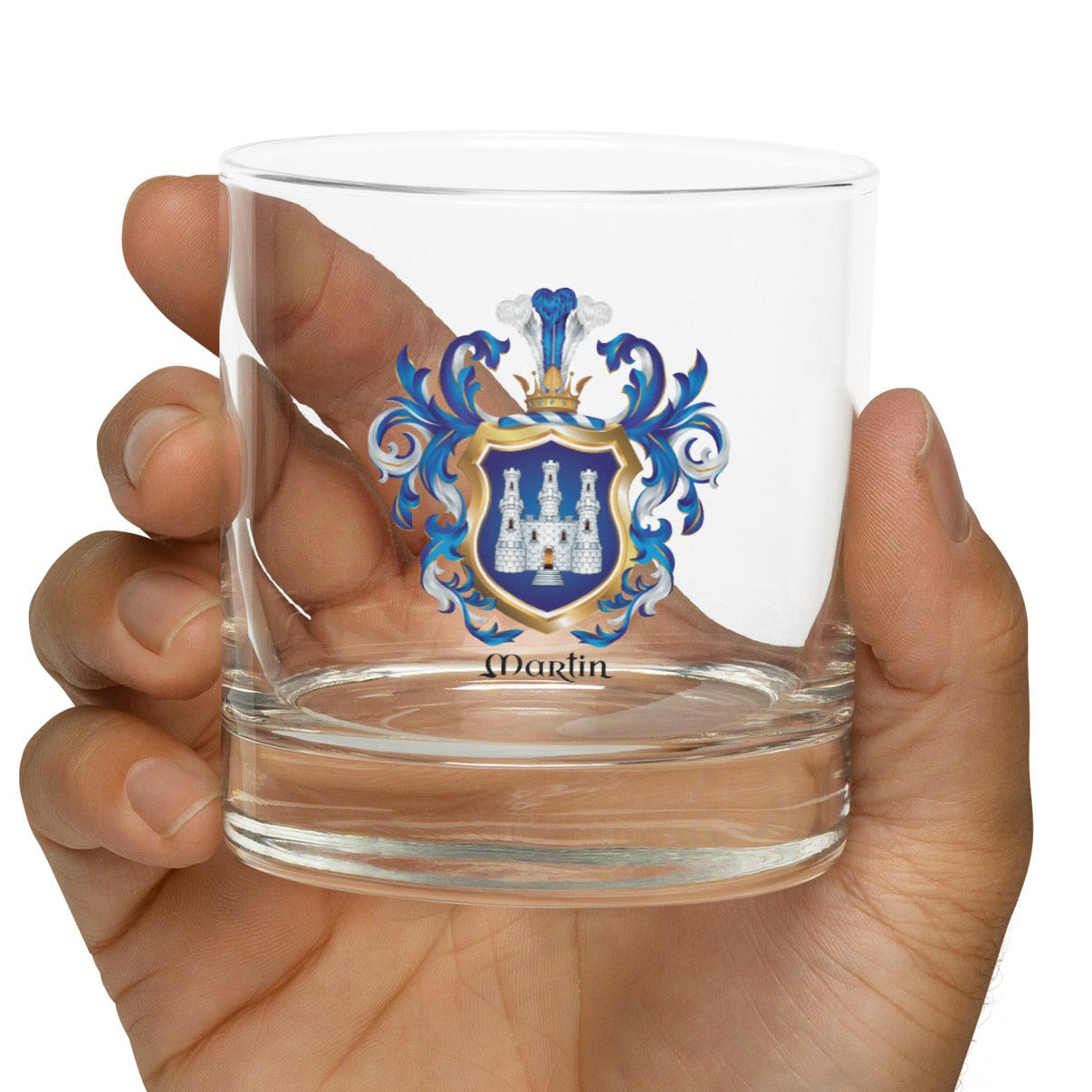Hand holding a clear glass with a Martin coat of arms with a white castle on a blue shield shown on a white background