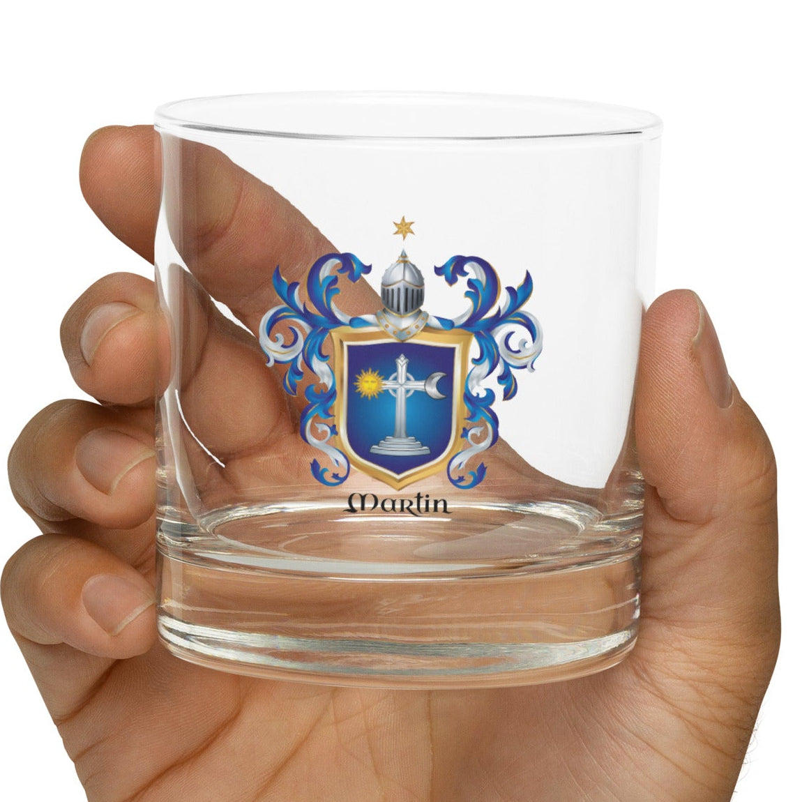 A hand holding a clear glass tumbler with a printed Irish Martin coat of arms blue and gold family crest and the name 'Martin' below it, against a plain white background.