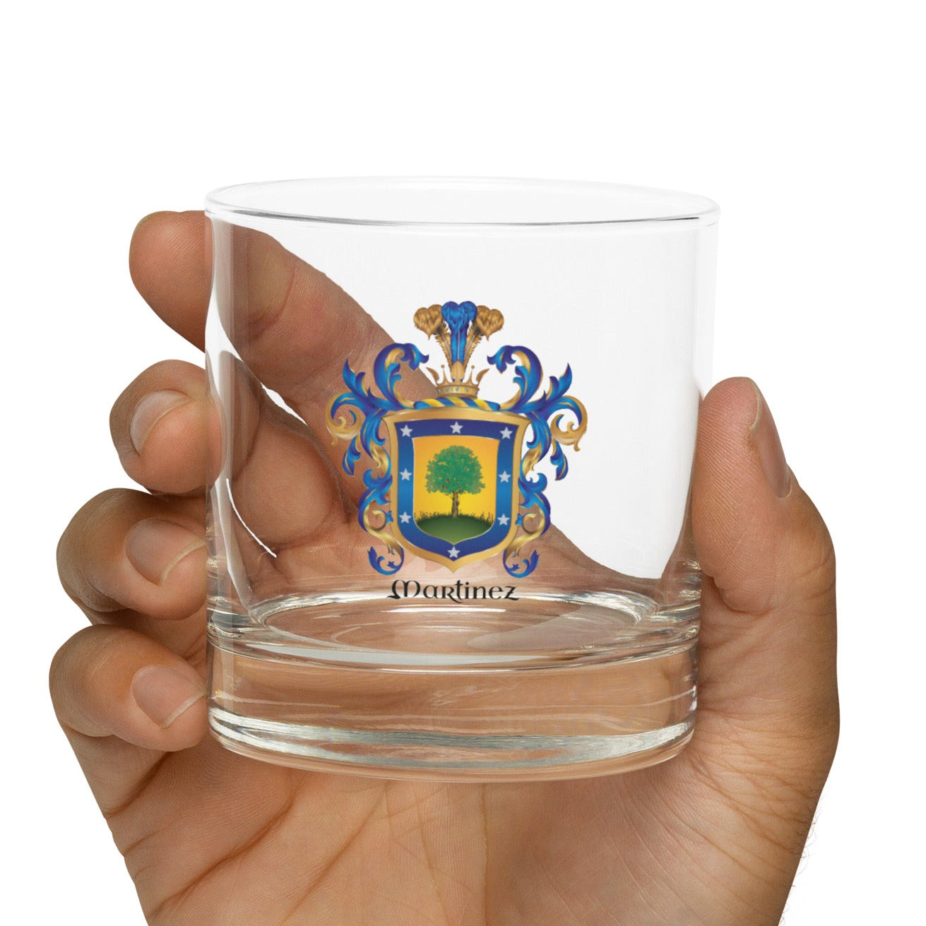 A hand holding a clear rocks glass tumbler with a colorful crest design and the name 'Martinez' printed near the base, against a plain white background.