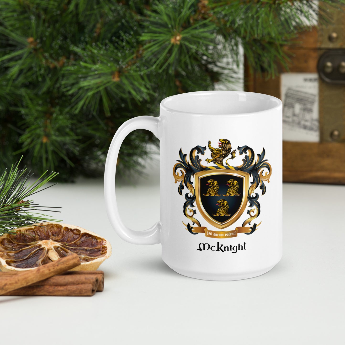 A white ceramic mug with a printed McKnight family crest and name, styled on a tabletop with pine branches, a dried orange slice, cinnamon sticks, and a wooden box in the background.