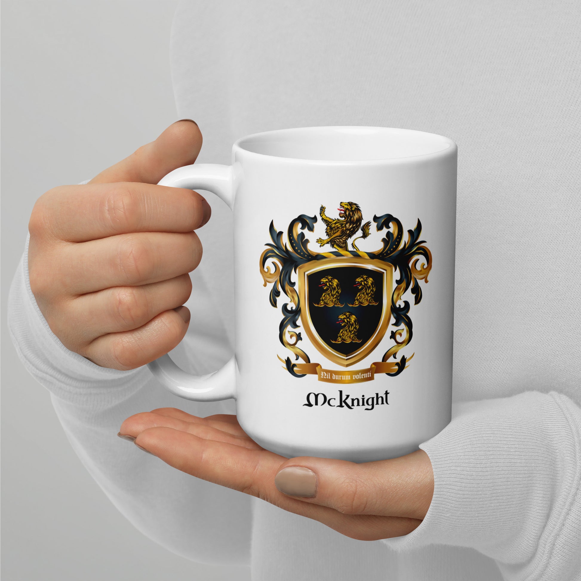 A white ceramic mug with a black, gold, and blue 'McKnight' family crest design, held by a person's hands wearing a white long-sleeve shirt against a light gray background.