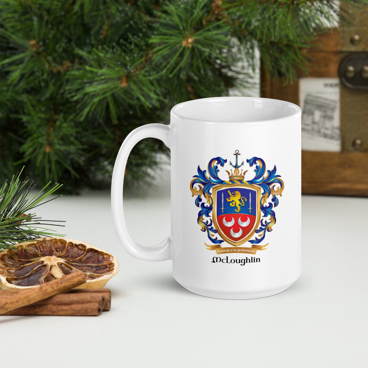 A white ceramic mug with a colorful McLoughlin family crest printed on the side, set on a tabletop with pine branches, a dried orange slice, cinnamon sticks, and a wooden chest in the background.
