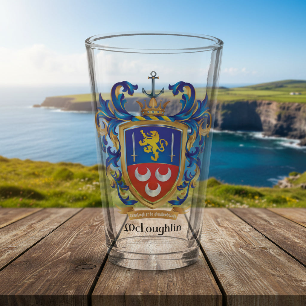 A clear pint glass featuring a printed McLoughlin family crest with a gold lion , two swords and 3 silver crescents and the name McLoughlin below amotto ribbon, shown on a wooden table top with a coastal Donegal  landscape in the background on a nice sunny day.