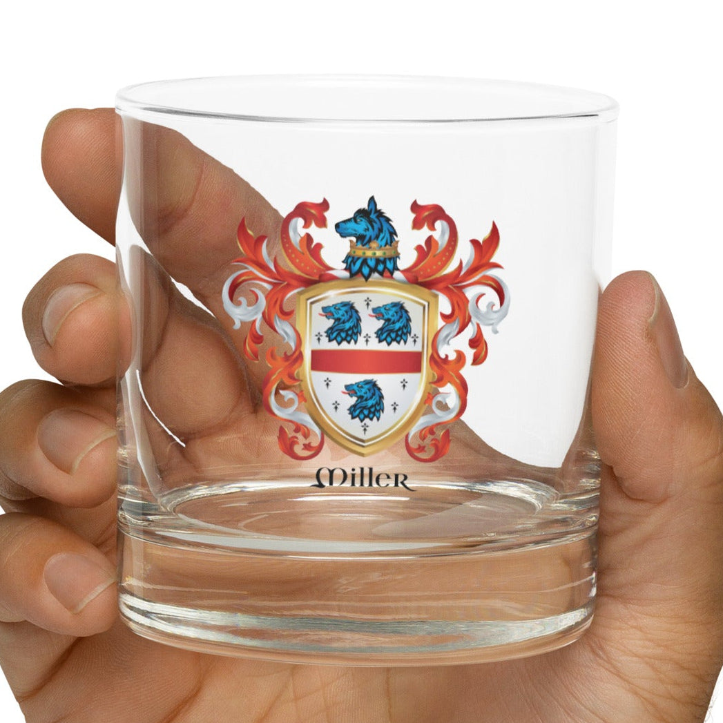 Hand holding a rocks glass with a Miller family crest coat of arms design on a white background