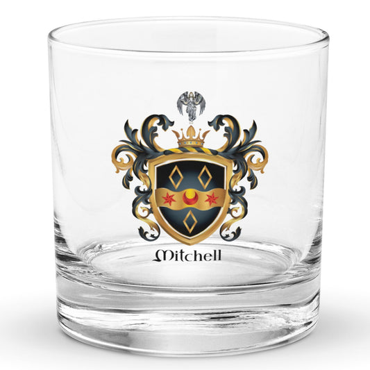 A clear classic heavy base rocks glass on a white background, printed with the Mitchell Family Crest featuring a black shield witha wavy gold ribbon with 2 red stars and a red cresent and 3 gold diamonds and an angel holding a dove above the shield on the crest.