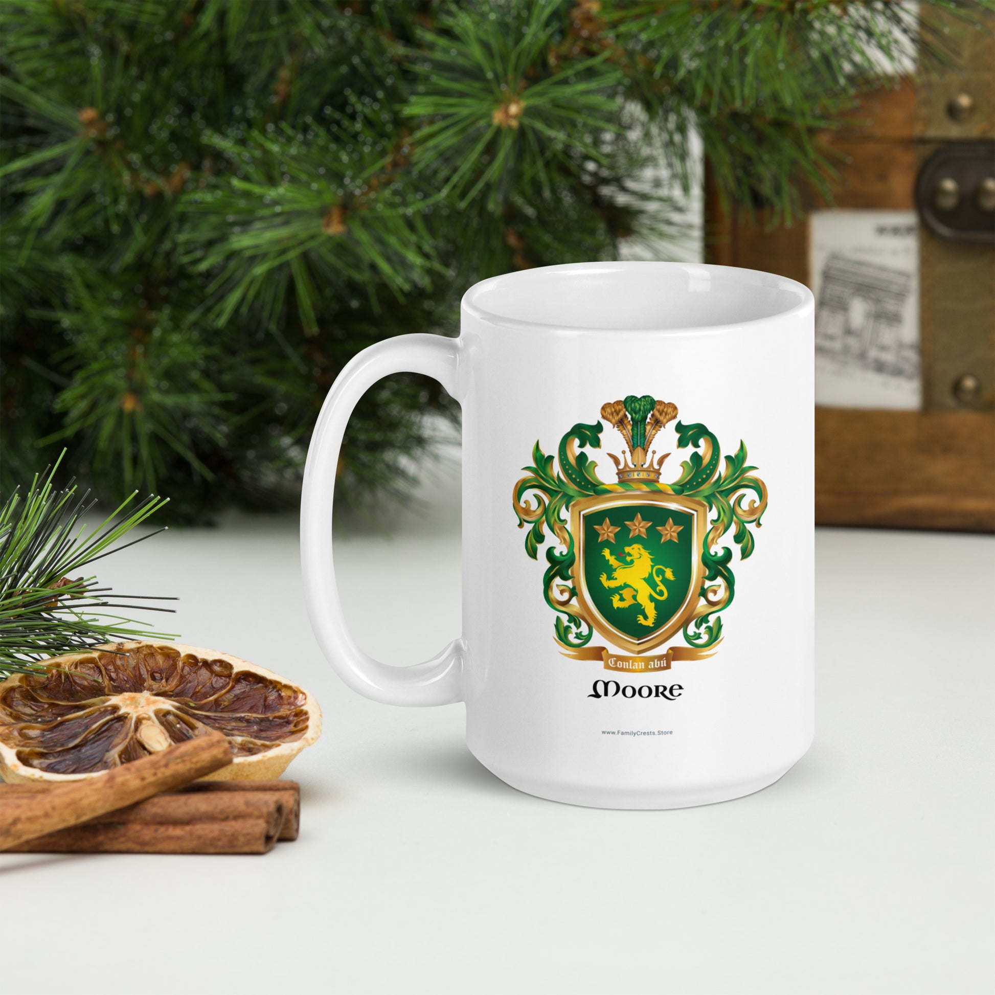 A white mug with the Moore Family Crest featuring a green shield with a golden lion and three gold stars with a crown on the shield and green and gold mantling all around with a motto'Conlan abu under the shield and the name Moore under the motto on a white surface with Christmas decorations.