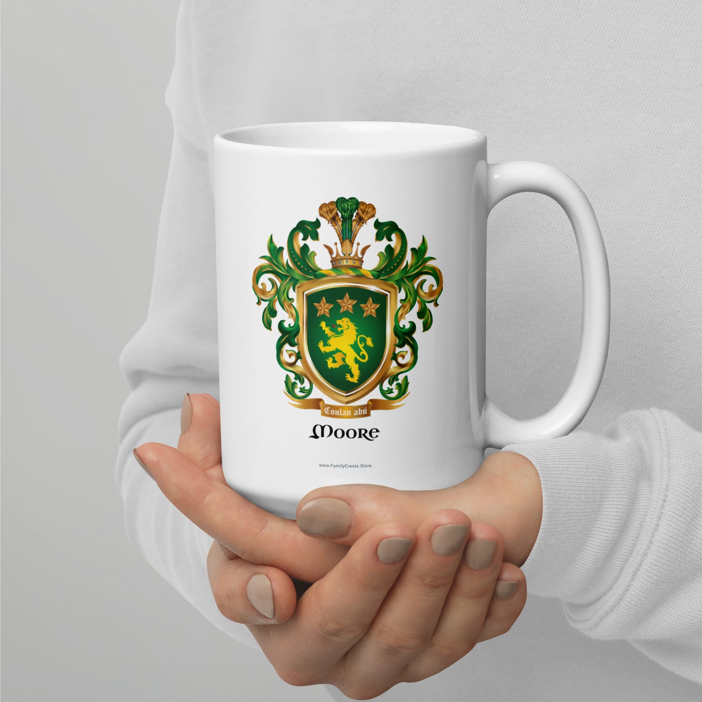 A white mug with the Moore Family Crest featuring a green shield with a golden lion and three gold stars with a crown on the shield and green and gold mantling all around with a motto'Conlan abu under the shield and the name Moore under the motto held by a person against a gray background