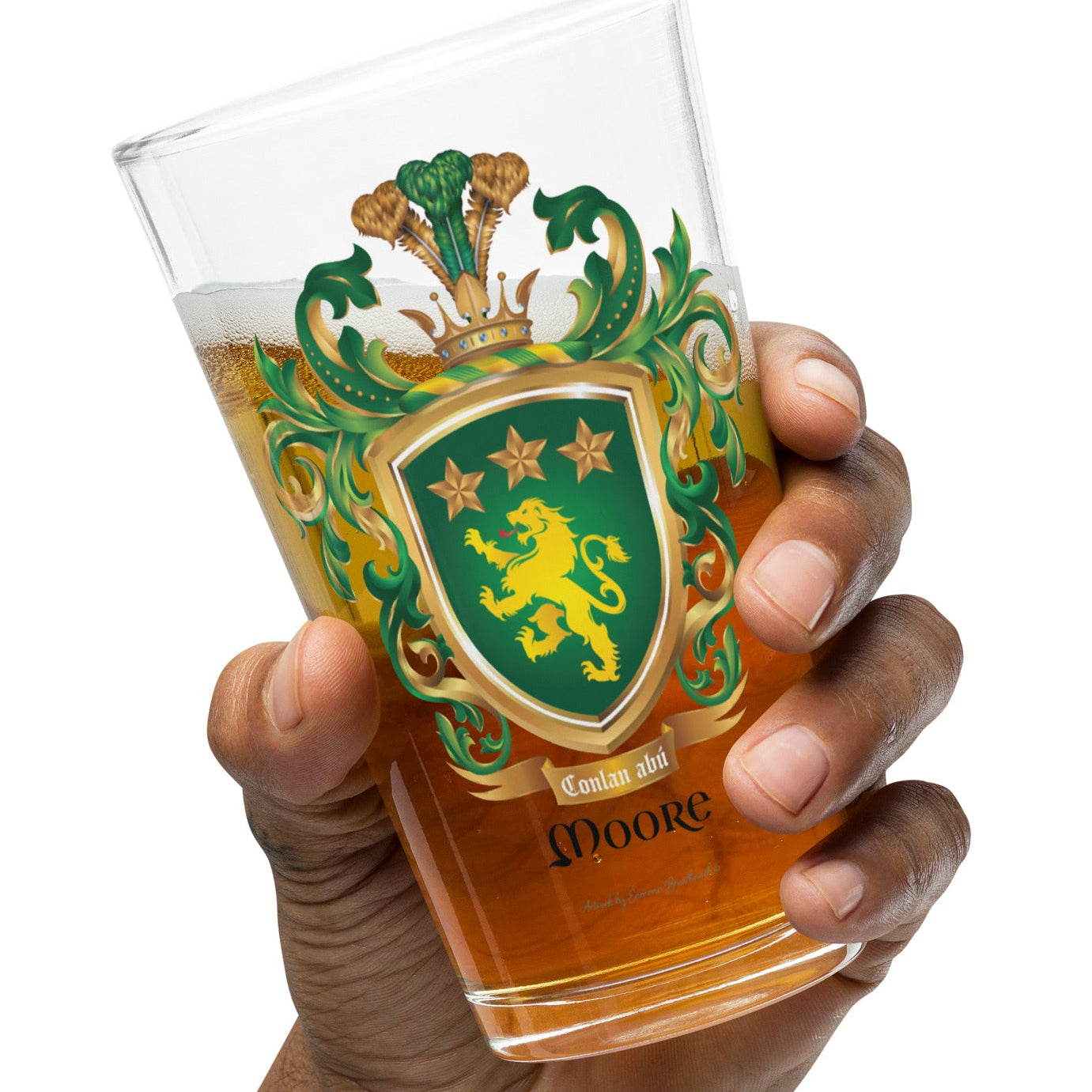 A hand holding a pint of beer in a pint beer glass featuring The Moore Family Crest showing a green shield with a gold lion and three gold stars above the lion with the motto and Moore name below the shield and a crown above and green and gold mantling all around  on a white background