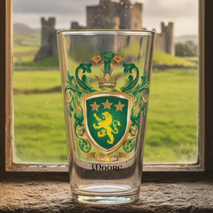 Clear pint beer glass featuring The Moore Family Crest showing a green shield with a gold lion and three gold stars above the lion with the motto and Moore name beloe the shield and a crown above and green and gold mantling all around  with the pint glass sitting on a window sill and a castle visible in the background through the window.
