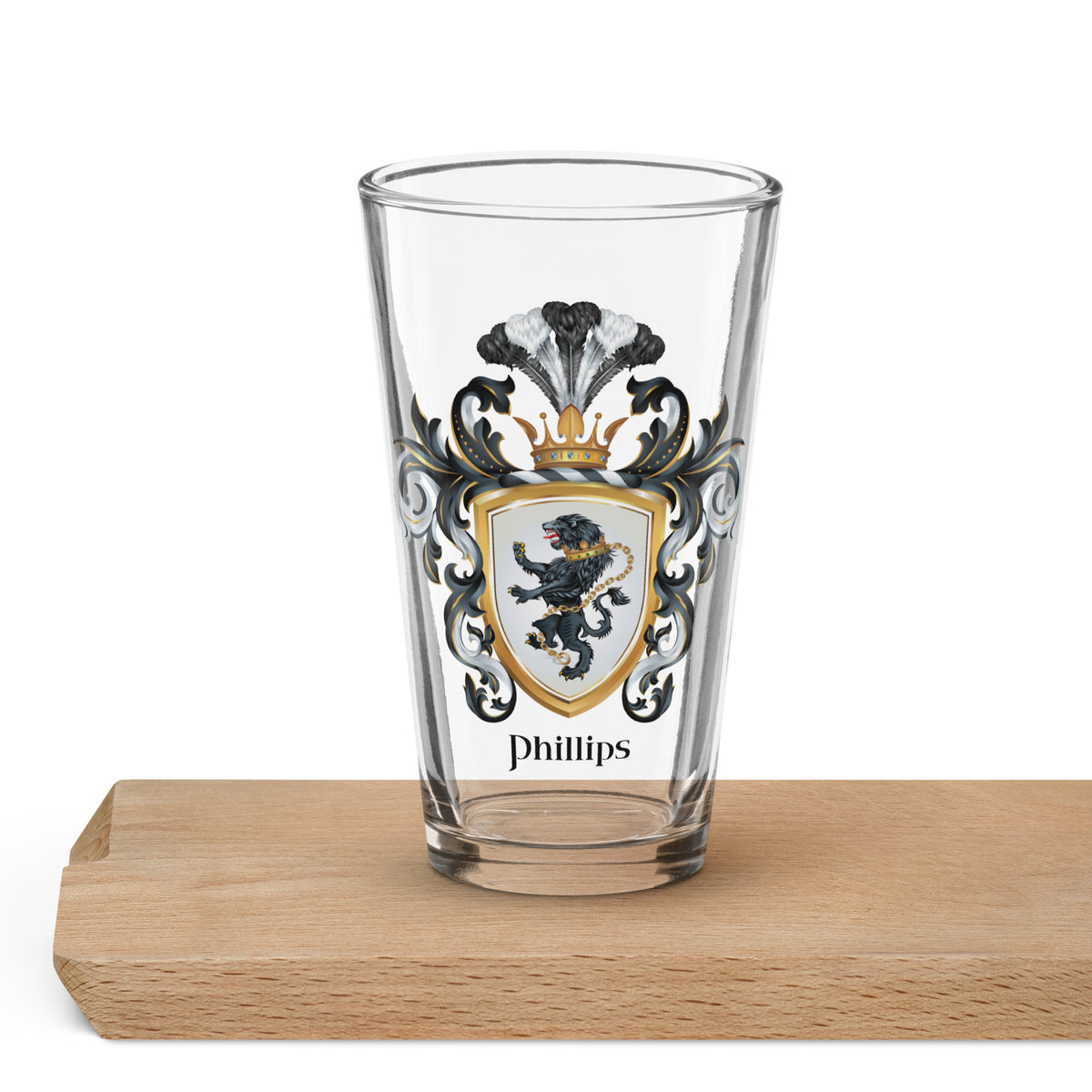 A clear pint glass with a custom family crest printed on the front, featuring a black and gold lion shield, ornate black scrollwork, and the name 'Phillips' below. The glass is shown standing on a light wood block against a white background.