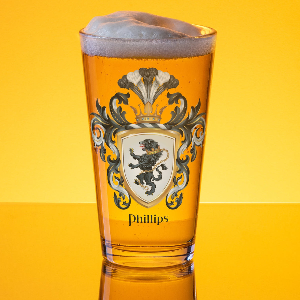A clear pint glass filled with golden beer and topped with white foam, featuring a printed crest with the name Phillips below, set against a bright yellow gradient background.