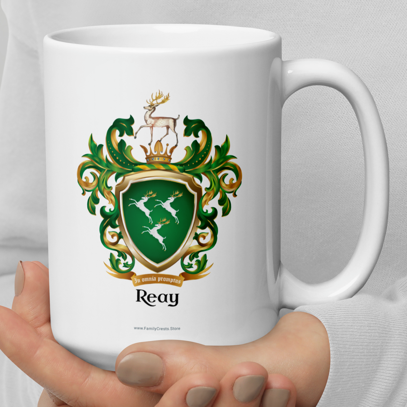 White mug with a green and gold crest Reay Family Crest featuring 3 white deer held by a person against a light gray background