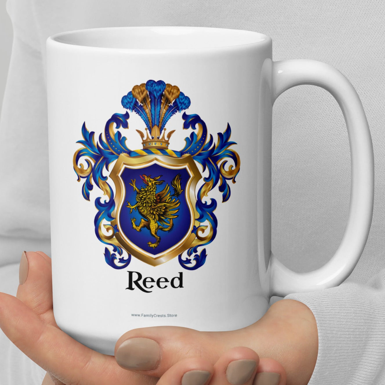White mug with a Reed Family Crest featuring a gold griffin on a blue shield and 'Reed' in black text held by a person against a light gray background