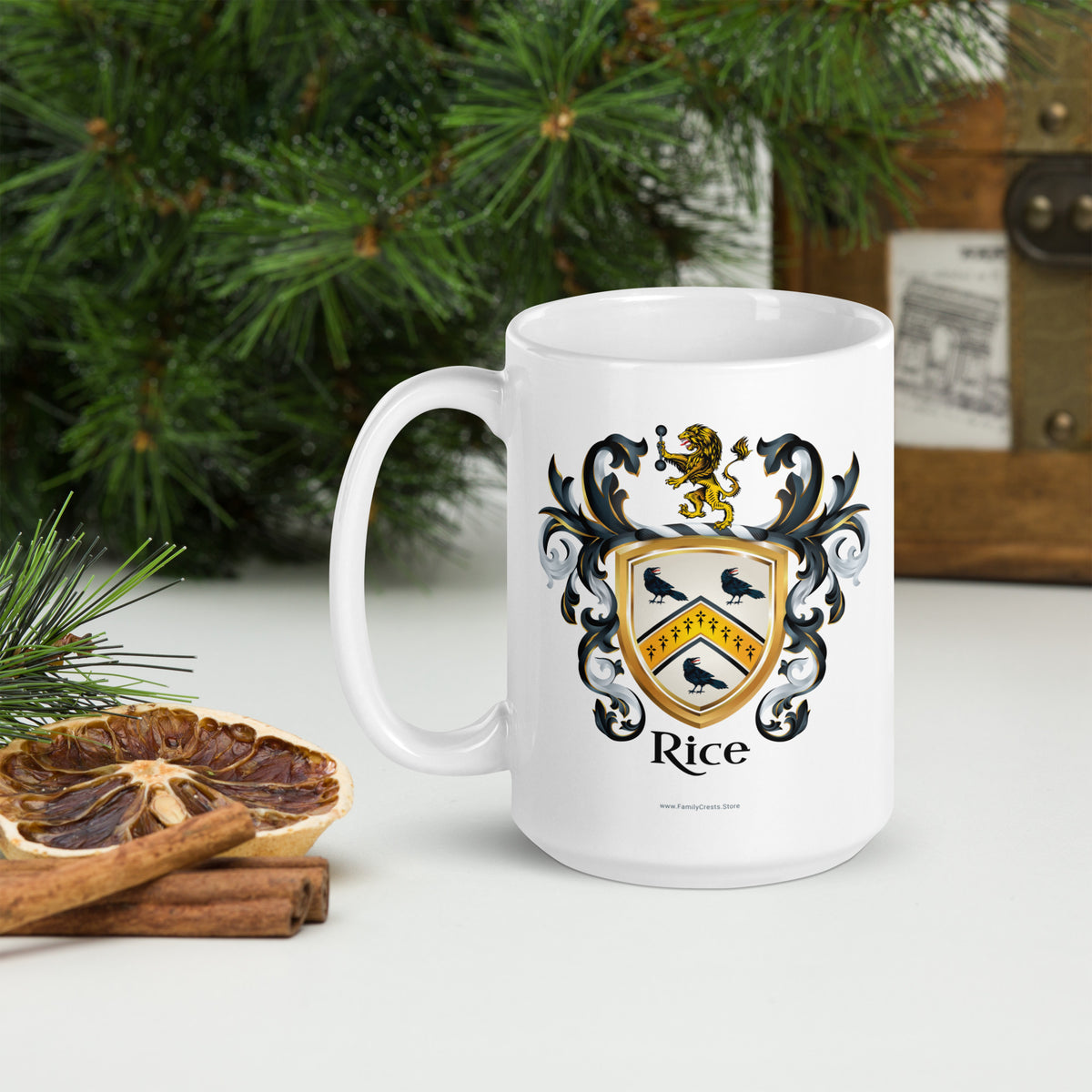 A white ceramic mug featuring a printed family crest with the name 'Rice' and black birds on a gold shield, styled on a tabletop with pine branches, cinnamon sticks, and a dried orange slice.