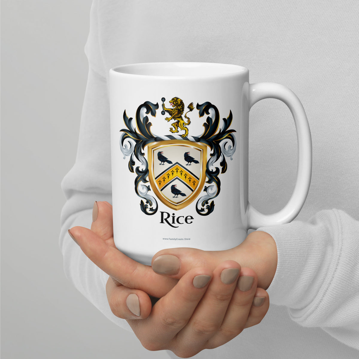 A white ceramic mug with a detailed family crest design featuring the name 'Rice' and a gold lion, held by a person's hands wearing a white long-sleeve shirt against a light gray background.