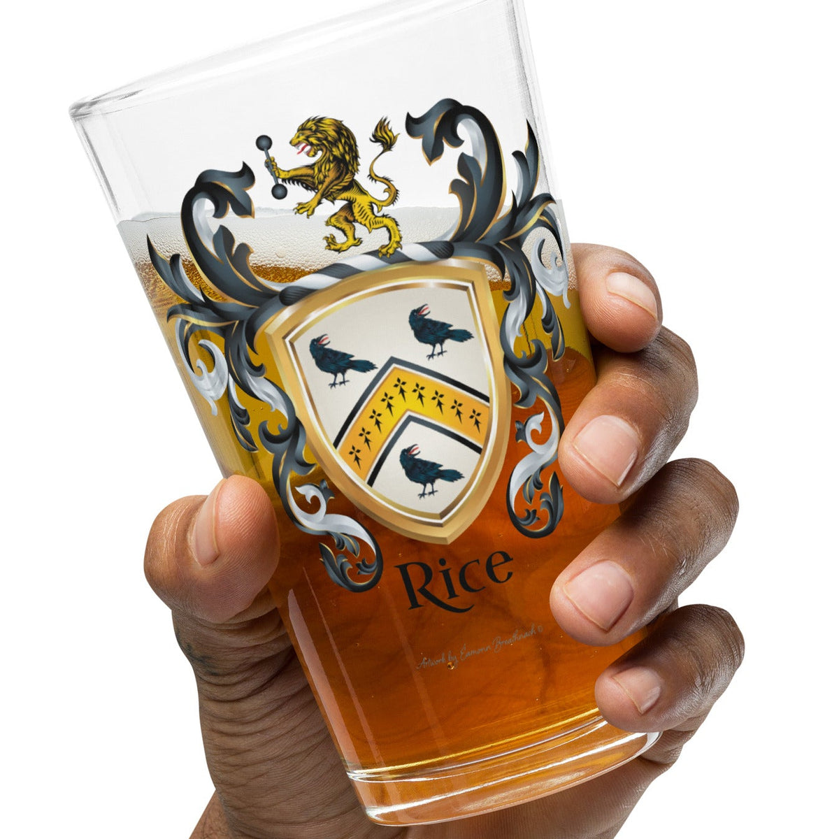 A hand holding a clear pint glass filled with amber beer, featuring a Rice Family Crest design and the word 'Rice' near the bottom, against a plain white background.